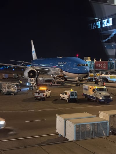 An aerial view captures a KLM Asia aircraft, a Boeing 777-300ER, parked at Aeropuerto Internacional Ezeiza, Argentina, at night. The aircraft, identified by its registration "PH-BVA," is a significant subject. In front of the aircraft, a Brinks armored truck, a white van, and a utility vehicle are positioned. A cargo loader is attached to the aircraft's belly. Baggage containers are visible on the tarmac. In the background, a jet bridge connects to the terminal, with visible signage including "ICBC" and a distorted "LA PAZ" sign, suggesting the terminal is branded as "La Paz." The scene is illuminated by the aircraft's lights, vehicle lights, and the ambient airport lighting, highlighting the aircraft's blue and white livery.