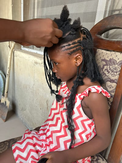 An individual's dark-skinned hands are observed braiding the natural, dark hair of a young Nigerian girl, approximately 5-8 years old, in Ashipa, Nigeria. The girl is depicted in profile, facing left, seated on a wooden chair with a patterned upholstered backrest. She wears a sleeveless top or dress featuring a red and white zigzag pattern with ruffled shoulder straps. Her hair is in various stages of braiding, with some sections formed into cornrows close to the scalp, others into loose individual braids, and remaining sections unbraided, forming tufts on the crown. The background features a textured, light-colored concrete or stucco wall on the left, against which a wooden-handled metal shovel is propped. A window with a fine mesh screen is visible behind the braider's hands in the upper background.
