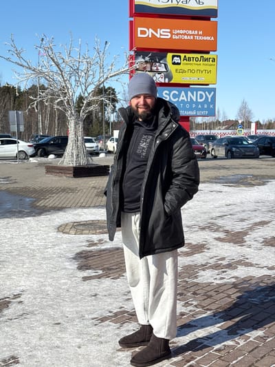 A man wearing a dark coat, light-colored sweatpants, a grey beanie, and brown shoes is standing in front of a sign with the names of various commercial establishments: "DNS - Digital and Household Appliances," "AutoLiga - Joint Trips, Let's go along the way," and "SCANDY-Topark - Retail Center." A decorative, lit-up tree is behind him, and a row of parked cars can be seen further back. The ground is covered in a mix of brick and snow. The scene is captured in Kogalym, Russia.