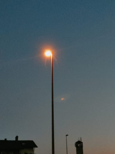 The image depicts an outdoor scene during twilight, possibly at dusk, with a gradient of colors in the sky transitioning from a clear blue at the top to a softer orange-grey near the horizon. A prominent streetlamp with a bright, warm orange glow is positioned in the center-right of the frame, casting light. In the lower portion of the image, silhouetted structures are visible. On the left, a dark-colored building with a pitched roof and a chimney is discernible. To the right of the building, a smaller, thinner streetlamp is present, and further to the right, a tower-like structure with a dark top is visible. The overall image has a grainy texture, suggesting it was taken with a camera in low light conditions. There are no people or specific actions depicted. The scene appears to be in an urban or suburban environment, given the presence of streetlights and buildings. The context of Murska Sobota, Slovenia, is provided, suggesting this is a location within that city.