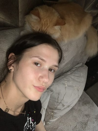 The image shows a close-up selfie of a young woman lying down, with an orange cat asleep behind her. The woman is looking towards the camera with a slight smile. She has dark hair and is wearing a black t-shirt and a gold necklace. The cat is nestled on a grey pillow. The background appears to be a sofa with textured pillows, suggesting an indoor setting. There is no discernible information within the image that would indicate a location in Belogorsk, Russia, or any other specific city.