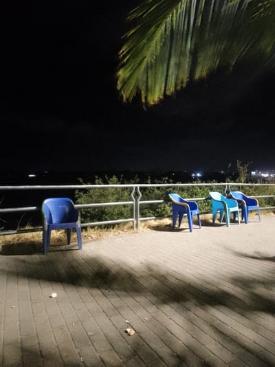 Four plastic chairs stand on a paved terrace overlooking dark greenery and distant lights at night. A metal railing borders the terrace. The sky is a deep black, partially obscured by the fronds of a large plant. The foreground of the terrace has several pieces of litter scattered across its surface, illuminated by an unseen light source.