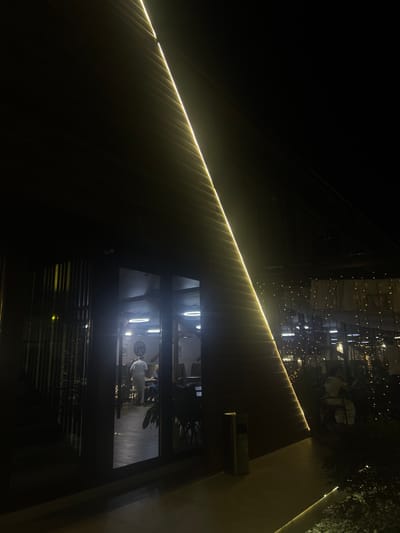 The image depicts a nighttime scene at the Achadara Restaurant in Achadara, Abkhazia. A striking illuminated architectural feature, a large angled surface with a continuous yellow light strip, dominates the left side of the frame. To the right, the interior of the restaurant is visible through large glass doors, revealing a warmly lit space with circular ceiling lights and tables. Several patrons are seated at tables, and a person in a white uniform stands near the entrance. Outside, decorative string lights twinkle amongst foliage. The overall ambiance is one of a modern establishment illuminated for evening operation.