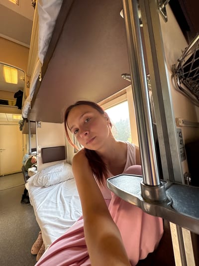 The image shows a young woman in a bunk bed compartment on a train. She is looking directly at the camera, with a slight smile. She is wearing a white tank top and pink pants. The compartment is small and appears to be a typical train sleeping car with bunk beds. There is a window to the right of the woman, showing trees and sunlight outside. The environment suggests travel within Russia, possibly on a long-distance train. The specific location of Sovkhoznyi, Russia is not explicitly indicated by any visual cues in the image.