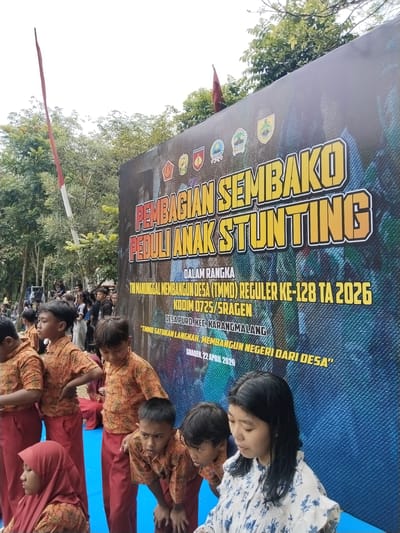 The image captures a scene outdoors, likely in the city of Puro, Indonesia, during an event focused on child stunting.

In the foreground, a group of children is visible, all wearing similar patterned orange and red outfits. One child wears a red hijab. A woman in a white and blue patterned blouse is also visible. She is possibly overseeing or involved in the event.

Behind the children, a large banner is prominently displayed with the words "PEMBAGIAN SEMBAKO PEDULI ANAK STUNTING" in large yellow letters. Other text on the banner reads "DALAM RANGKA TNI MANUNGGAL MEMBANGUN DESA (TMMD) REGULER KE-128 TA 2026 KODIM 0725/SRAGEN DESA PURO, KEC KARANGMALANG TMMD SATUKAN LANGKAH, MEMBANGUN NEGERI DARI DESA" and "SRAGEN, 22 APRIL 2026". The banner includes the Diponegoro seal and what appear to be other insignia.

In the background, more people are gathered. A red flag is visible. Trees are also noticeable in the background. The scene suggests a community initiative or campaign addressing child stunting issues.