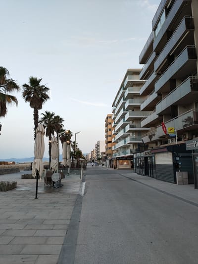 The image depicts a promenade in Canet-en-Roussillon, France, likely in the late afternoon or early evening given the lighting conditions. On the right side of the image, a modern multi-story building with numerous balconies dominates the scene. Several shops with their shutters down are visible at the ground level of this building, suggesting it might be off-season or nearing closing time. Some of these shops appear to be restaurants or bars. Above, on the upper floors, balconies are visible, some with modern glass railings.

To the left of the promenade, palm trees are planted in a row, with some umbrella-covered tables and chairs stacked nearby, indicating a seasonal outdoor seating area. Further to the left, a beach and the sea are partially visible, with a low wall and wooden fencing separating the promenade from the sand. In the distance, a range of hills or mountains can be seen. The sky is clear with a few wispy clouds, and a small airplane is visible in the far distance.

A few individuals are seen walking along the promenade in the middle ground, suggesting the area is accessible but not crowded. The promenade itself is paved with large rectangular stones, and a darker strip runs down its center. The overall atmosphere is calm and peaceful, characteristic of a seaside resort.