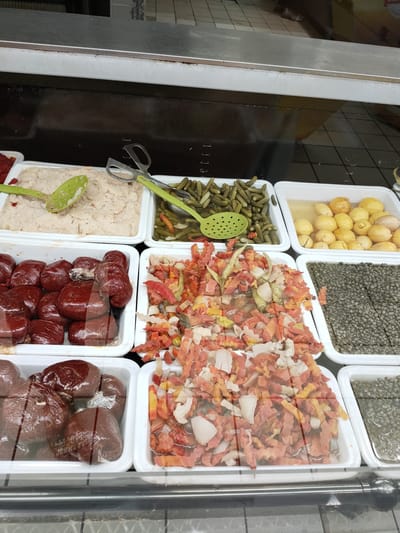 In a brightly lit refrigerated display case, a variety of preserved foods are presented in white rectangular trays. The items include a creamy, pale spread in the top left, likely a dip or pâté, served with a green slotted spoon. Next to it are small, pickled gherkins in brine, also with a green slotted spoon and a pair of metal tongs. To the right of the gherkins are small, whole lemons, preserved in a light yellow liquid.

Below these, in the front row, are several trays of chopped and mixed ingredients. The leftmost tray contains what appear to be small, dark red, individually wrapped food items, possibly sausages or meat preparations. Adjacent to this is a tray filled with a colorful mix of chopped vegetables, predominantly orange and white, with hints of red and green, possibly a salad or relish. To the right of the vegetable mix, another tray holds small, dark green, round objects, likely capers, in brine. Finally, in the bottom right, a tray contains more of these capers, with some other preserved items visible. The overall scene suggests a deli counter or a section of a grocery store specializing in preserved goods. There are no people visible in the frame. The lighting appears to be artificial, typical of an indoor commercial setting.