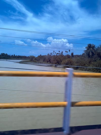The image shows a wide, muddy river flowing through a tropical landscape under a bright blue sky with scattered white clouds. The riverbanks are densely covered with lush green vegetation, including numerous palm trees. In the foreground, a bright yellow railing runs horizontally across the lower part of the frame, obscuring the view slightly. Several dark electrical wires are strung across the upper portion of the image, appearing as thin black lines against the sky. The scene appears to be a view from a bridge or elevated structure, possibly overlooking a part of the city of Padangrubek, Indonesia, known for its rivers and tropical flora.