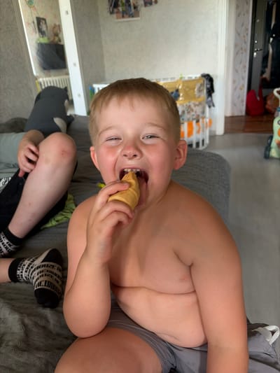 A boy is seen in a close-up shot, with his torso and head filling most of the frame. He is shirtless and wearing gray shorts. The boy is holding an ice cream cone up to his mouth and appears to be taking a bite, with his mouth open and eyes looking towards the camera. His expression suggests he is enjoying the treat. He has light brown hair and fair skin.
In the background, part of a couch with a gray textured fabric is visible. The scene appears to be indoors, possibly in a living room or bedroom, given the presence of furniture and what looks like a shelf with various items. The lighting suggests it might be daytime, but there are no clear indicators of the time of day.
Below the boy's torso, another person's legs are visible, wearing black shorts and black and white patterned socks. One of the socks has visible text in black lettering against a white background, which reads "LONDON." The legs are bent, with the person sitting or reclining.
The overall mood of the image is casual and domestic, capturing a candid moment of a child enjoying a snack. The setting appears to be in Russia, given the location context.