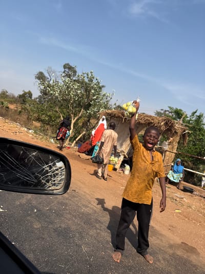 This image depicts an outdoor roadside scene in Nandu, Nigeria, under bright daylight with a clear blue sky.
In the foreground, the shattered surface of a vehicle's black side mirror is visible on the left, reflecting distorted elements of the environment. Directly in front of the mirror, a section of asphalt road stretches across the lower portion of the frame.
To the right of the asphalt, a young male, approximately 8-12 years old, stands barefoot on a dirt path. He is wearing a mustard yellow short-sleeved tunic and dark trousers. His mouth is open in an excited expression, and his right arm is raised high, holding a clear plastic bag containing several small, light green fruits, possibly limes or guavas.
Behind this boy and slightly to the left, an adult male walks away from the camera, heading towards the left. He wears a light tan tunic and trousers, carrying a black bag in his left hand and a bundle of red and white fabric over his left shoulder. Adjacent to him is a rustic, makeshift market stall or shelter constructed from wooden poles and a thatched roof, displaying various fruits and items on a low surface.
Further to the left, another adult, likely female, is partially visible from behind, walking away. She wears a dark top and a colorful patterned wrapper skirt, appearing to carry items.
In the mid-ground, a second thatched structure is visible. To the far right, seated next to this structure, an adult female is present, wearing a light blue head covering and a patterned blue and white dress.
The background consists of scrubby vegetation, trees, and a dirt slope, with scattered debris visible along the path. Strong shadows are cast on the ground, indicating a high sun position.