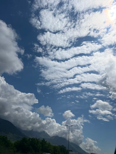 The image captures a vibrant sky filled with diverse cloud formations above a mountainous landscape, set in Kavač, Montenegro. The scene is dominated by a clear blue sky, partially obscured by clouds.

In the upper and left portions of the sky, delicate, elongated clouds stretch in parallel, rippled bands, reminiscent of altocumulus undulatus or cirrocumulus, with hints of a sun halo or glint in the upper left corner. These clouds appear lighter and more dispersed. Contrasting with these, large, fluffy cumulus clouds with significant vertical development are concentrated towards the right and bottom right of the frame, casting soft shadows upon themselves.

Below the expansive sky, the dark, forested slopes of mountains or large hills rise on the right side, partially enveloped or overshadowed by the larger cloud masses. Green tree foliage is visible at the very bottom right, at the base of these mountains.

A tall, dark streetlamp with two lights stands prominently on the right side, extending upwards into the sky. In the far background on the right, near the mountains, some indistinct man-made structures or signs are faintly visible, though no specific text is discernible.

The time of day appears to be daytime, likely mid-morning or afternoon, suggested by the bright natural light and the clear contrast between light and shadow. The weather is bright but very cloudy, with a mix of high-level and mid-level clouds. The overall mood is one of natural beauty and expansive calm.