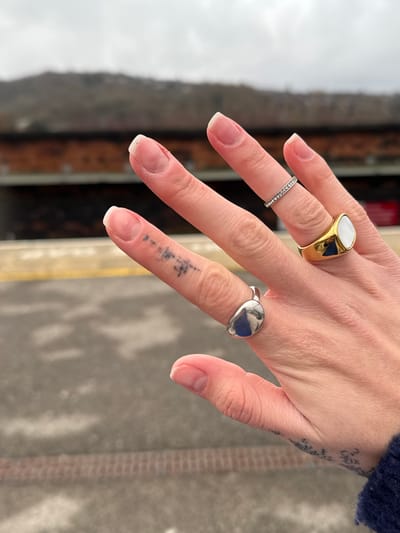 A hand is displayed with three rings. The index finger has a gold signet ring with a white stone. The middle finger has a thin silver band. The ring finger has a silver ring with a blue stone. The hand is pale with visible veins. There is a tattoo of vertical lines on the side of the middle finger, and another tattoo on the side of the hand, near the wrist. The nails are neatly shaped and have a natural look.