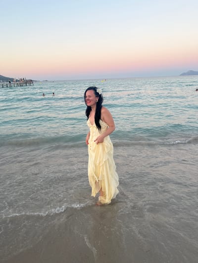 A woman with long black hair, wearing a flowing, pale yellow dress adorned with a white flower in her hair, stands knee-deep in shallow water at the beach. She is smiling and looking towards the camera, with her hand resting on her abdomen. The water is calm with gentle ripples, and the sandy seabed is visible. In the background, the sea stretches to the horizon under a soft, colorful sky, with hues of pink, orange, and blue suggesting either sunrise or sunset. A wooden pier with people on it extends into the water in the distance, and some other individuals are visible swimming or wading in the sea. The overall mood is serene and pleasant, capturing a moment of relaxation and beauty at the beach.
