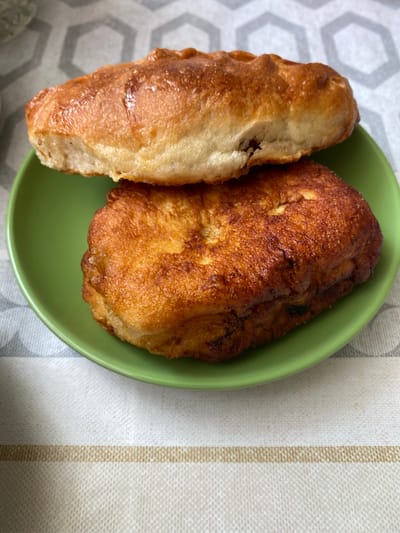 A close-up, overhead shot shows two baked goods, likely pastries, on a bright green plate. The pastry on top is golden brown with a slightly glossy sheen, and has a crimped edge. The pastry below it is darker, with a more textured, fried appearance. Both pastries are oval-shaped and appear to be resting on a patterned tablecloth. The background is blurred, with a grey and white hexagonal pattern visible. The image does not contain any information about the location in Novonikolsk, Russia.