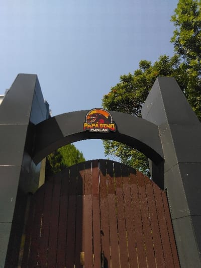 The image displays a dark grey archway structure positioned above a dark brown, vertically paneled wooden gate, viewed from a low-angle perspective. Centrally affixed to the arch is a circular emblem featuring a dinosaur silhouette against an orange and black background, with the white text "PAPA DINO PUNCAK" inscribed below the silhouette. Green tree foliage is visible through the arch opening and to the upper right of the frame, set against a clear blue sky. No individuals or active interactions are present. The scene is situated in Ban Xieng Lom, Laos.