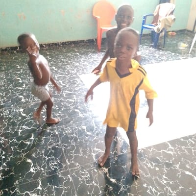 Three young boys are standing on a black and white tiled floor. One boy is wearing a yellow and black shirt and standing in the center of the image. He is facing the camera, smiling. The boy to his left is wearing blue and white striped shorts and appears to be giving a peace sign. The boy to the right of the yellow shirt boy is shirtless, wearing blue jean shorts, and is smiling. He is standing with his back to the camera. There is a bright orange plastic chair and a blue plastic chair in the background. The room has blue walls. The coordinates given are located near the city of Warri, Nigeria. The location provided is not the city of Adanisievbe.