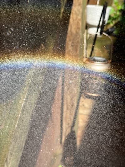 The image depicts a close-up view of water droplets suspended in the air, creating a rainbow effect. The light source is positioned in a manner that allows the dispersion of light into its constituent colors. The background appears to be an outdoor setting, with indistinct objects suggesting a garden or yard with a metal water pipe and faucet being the focus of the image. A white container, possibly a bucket, is visible in the background. The image was taken in Port Washington, USA.