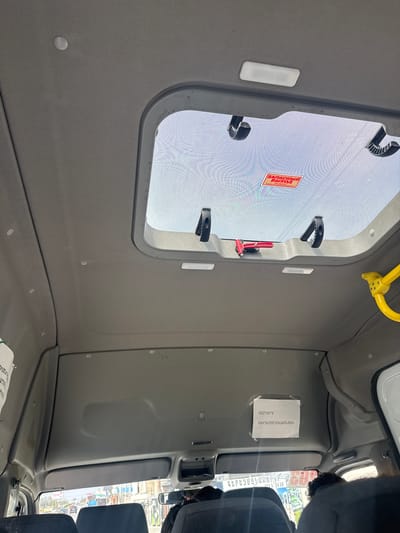 The image captures the interior of what appears to be a bus or a large van, viewed from a low angle looking upwards and slightly towards the front. The dominant feature is a rectangular roof hatch, through which bright daylight is visible, suggesting it's daytime. The hatch is surrounded by a gray frame and features several black latches, with a red emergency release mechanism prominently visible.
The interior ceiling and upper walls are covered in a uniform gray, textured fabric. A yellow grab handle or safety bar is mounted near the front right corner of the hatch. Towards the right side of the image, parts of dark fabric seats are visible, indicating the seating area of the vehicle. Through the front and side windows, a blurred outdoor scene of what looks like a street or city environment can be vaguely discerned.
Visible text provides specific details:
1. On the red sticker attached to the hatch, Russian text reads "ЗАПАСНЫЙ ВЫХОД" (EMERGENCY EXIT). Below it, smaller Russian text states: "ПОТЯНИТЕ РЫЧАГ ВВЕРХ И ОТКРОЙТЕ ЛЮК." (PULL THE LEVER UP AND OPEN THE HATCH.)
2. A white paper sign taped to the ceiling/wall on the right displays Armenian text. The main phrase is "ԱՐԳԵԼՎՈՒՄ Է ԾԽԵԼ" (ARGELVUM E TSXKHEL), which translates to "SMOKING IS FORBIDDEN."
3. Below this sign, partially visible text on an exterior sticker or through a window contains Armenian words like "ԳԵՐԵԶՄԱՆԱՊԱՏ" (GEREZMANAPAT), meaning "CEMETERY" or "GRAVEYARD," along with numbers like "1430," possibly indicating route information or a contact number.
The interior appears clean and functional, typical of public transport or a commercial vehicle in Ptghunk, Armenia.