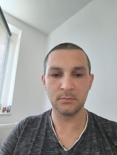 This is a medium close-up selfie of a man with a buzz cut and light stubble on his face. He is wearing a dark gray t-shirt with thin lighter gray horizontal stripes. The t-shirt has a light-colored trim around the V-neck. He is looking directly at the camera with a neutral expression.

The background is a plain white wall. To the left, there is a window with horizontal blinds. Below the window, a white radiator is visible. On the windowsill, to the left of the window, are three small framed pictures. The lighting suggests it is daytime.

The location context is Wolgast, Germany. There is no visible text in the image.