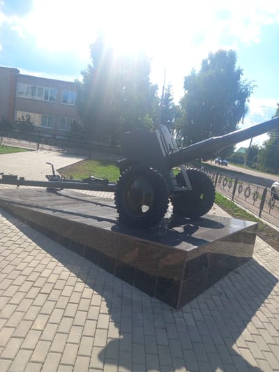 The image depicts an outdoor scene with a monument to a cannon situated on a paved area. The monument rests on a dark, polished stone base. The cannon is dark green and features two large wheels. To the left of the cannon, a portion of another similar structure is visible. The background includes a brick building with multiple windows, trees, and a street with a fence. The sun is bright, casting shadows and creating lens flare effects. The location is identified as Sterlibashevo, Russia.
