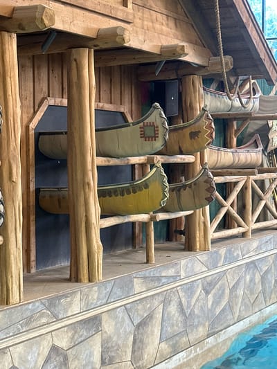 A decorative display of canoes is mounted on a wooden structure, suggesting an indoor or covered outdoor environment with a rustic theme. The canoes, varying in color and design, are arranged on multiple shelves supported by thick, natural wood posts and beams. Some canoes feature decorative patterns, including what appears to be a geometric design on one and a bear silhouette on another.
The background is a dark, textured wall, likely part of the building's interior. Below the canoes, a multi-layered structure with a stone-like pattern is visible, leading down to a body of water with a rippling surface, indicating a pool or water feature. A rope is suspended from the upper wooden structure, adding to the nautical or wilderness aesthetic. The overall scene is well-lit, highlighting the textures of the wood and the details of the canoes. The location is identified as Centralia, United States of America.