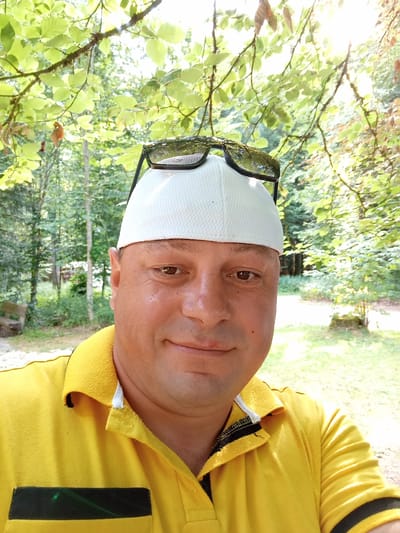 A medium shot photograph captures a fair-skinned adult male wearing a yellow polo shirt and a white baseball cap. Sunglasses are perched on top of the cap. The man is looking slightly to his right with a slight smile. The background suggests an outdoor setting with lush green foliage and trees, indicative of a park or forest area. Sunlight filters through the leaves, creating a dappled effect on the scene. The location is Meiniswil, Switzerland.