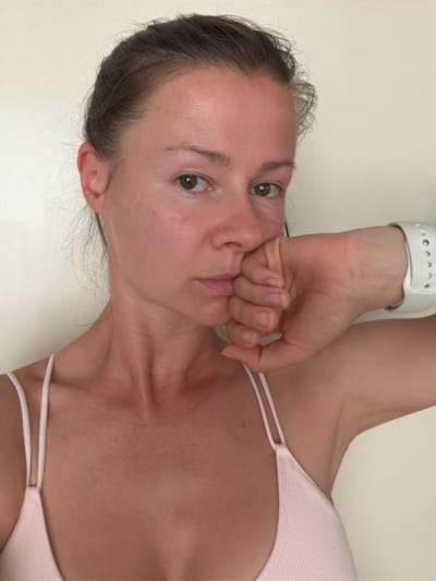 The image shows a close-up of a woman's face and upper body. She has her hair pulled back, and her left hand is resting on her mouth. She is wearing a light pink camisole. A white smartwatch is visible on her left wrist. The background is a plain, light-colored wall. There is no information in the image to suggest it is located in Melick, Netherlands.