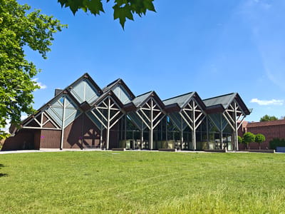The image depicts a modern building with a distinctive angular roof design, set against a clear blue sky on a sunny day. The building features large glass windows interspersed with wooden and light-colored framing, giving it an open and airy appearance. The roof is dark and sloped, with a series of peaks that create a dramatic silhouette.
In the foreground, a lush green lawn stretches out, with some areas of taller, wilder grass. To the left of the building, a large tree with vibrant green leaves partially overhangs the scene, casting dappled shadows. The overall impression is one of tranquility and architectural interest.
The building's exterior is characterized by its repeated triangular elements, both in the roof structure and the framing of the glass sections. The wooden facade on the left side of the building is darker and more solid, contrasting with the transparency of the glass-fronted sections. Two circular signs with red borders and a diagonal line are visible on the wooden facade, suggesting a prohibition of some kind, possibly related to parking or access.
Behind the main building, a red brick structure is visible, along with some neatly trimmed trees. A person can be seen in the distance, near the brick building, adding a sense of scale to the scene. The bright sunlight indicates it is daytime, likely late morning or afternoon, with no visible clouds except for a few wisps high in the sky. The location is identified as Memmingen, Germany.