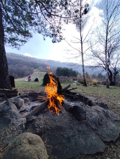 An outdoor scene in Маруцековци, Bulgaria, depicts a campsite or recreation area under daylight. In the immediate foreground, a wood campfire burns brightly with orange and yellow flames within a circular fire pit constructed from large, dark grey stones. Charred wood and ash are present in and around the burning logs. To the left, a portion of a thick tree trunk is visible. In the midground, approximately 15 meters from the fire, two individuals are seated on a grassy slope. One person, wearing a pink top, is clearly visible reclining, while the second individual is partially obscured. Behind them, a small body of water is visible, bordered by a line of evergreen trees. Beyond the water, a cluster of bare-branched deciduous trees, possibly birch, stands against the sky. The background features a range of hills or mountains covered with sparse vegetation and bare trees under a bright sky with scattered clouds. The upper left of the frame is partially obscured by the dark needles and branches of an evergreen tree.