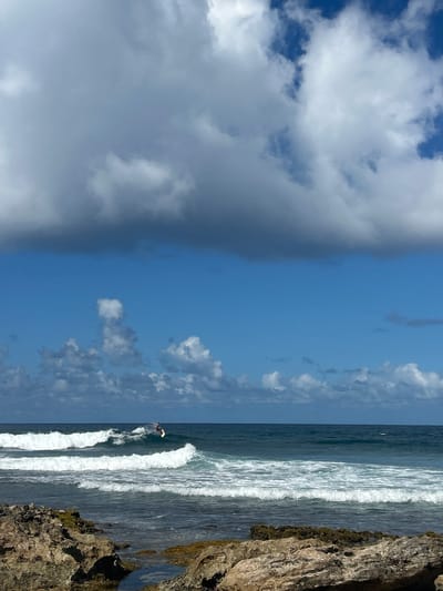 The image captures a coastal scene in Le Moule, France, featuring a surfer on a wave under a partly cloudy sky. The upper half of the frame shows a blue sky with varying cloud formations; a large, dense grey cloud bank occupies the upper left quadrant, while smaller, whiter cumuliform clouds are scattered towards the right and along the horizon. The mid-ground displays the open ocean, characterized by deep blue and green water with visible wave patterns. A single individual, identified as a surfer, is positioned on a breaking wave towards the center-right of the oceanic expanse, standing on a light-colored surfboard with water spray visible around them. Another wave, further in the foreground, is also breaking. The lower portion of the image reveals a rocky shoreline composed of irregular, light brown and grey rock formations, interspersed with small tidal pools and patches of dark green marine growth.