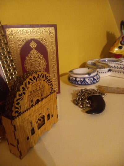 A decorative book with a gold, filigree border and burgundy center sits alongside an ornately carved wooden box. Behind them hang a collection of ornaments, including glazed ceramic pieces in white and blue, and a dark, shiny ornament attached to a silver chain. The background is a blend of yellow and light beige.