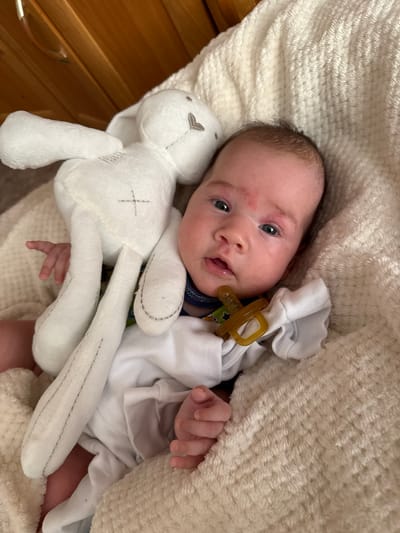 A newborn baby is lying on a cream-colored fluffy blanket. The baby is wearing a white onesie and has a tan pacifier in its mouth. To the left of the baby is a white stuffed bunny. The background shows wooden furniture. The scene is indoors.