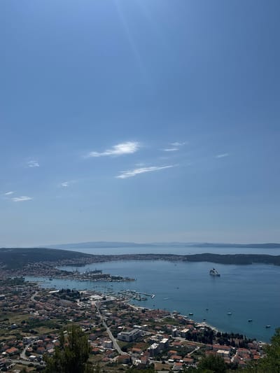 The image displays a panoramic view of a coastal city, identified as Seget Donji, Croatia. The scene is captured from an elevated vantage point, looking out over a bay filled with boats of various sizes, including sailboats and a large cruise ship positioned further out at sea. The foreground features dense vegetation and trees, hinting at a hillside perspective. Below, the city unfolds with numerous red-tiled roofs, characteristic of Mediterranean architecture, interspersed with modern buildings. A prominent harbor area with industrial cranes and docked vessels is visible along the coastline. The water is a deep blue, reflecting the clear sky above, which is dotted with a few wispy white clouds. The background shows rolling hills and distant islands, creating a sense of depth and scale. The overall impression is one of a vibrant, active coastal community under a bright, sunny sky.