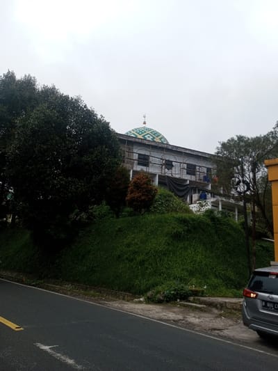 A building with a green and yellow patterned dome is under construction. Scaffolding is visible around the structure. Lush green vegetation covers the hillside in front of the building, with a large tree obscuring part of the view. A paved road is in the foreground, with a parked silver vehicle partially visible on the right side of the frame. The sky is overcast and gray.