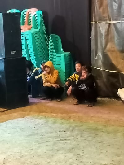 The image captures a scene in what appears to be a semi-enclosed or temporary event space, likely at night or in low light, in Kecamatan Cimaung, Indonesia. In the foreground, several young men are squatting or sitting on the ground, facing generally towards the right (out of frame).

There are at least four young men visible. The one furthest to the left is partially obscured by two large, black, rectangular speakers stacked vertically. The next young man is wearing a yellow hooded sweatshirt (hood up) and dark pants, and he is squatting with his hands near his mouth. To his right, another young man, also wearing a yellow top with black accents, is looking towards the right. The person furthest right among the group has dark hair and is wearing a black hooded sweatshirt with white patterns on the front, also squatting. All of them appear to be wearing casual attire, and some are wearing sandals.

In the background, a large number of bright green plastic chairs are stacked neatly in several columns, suggesting preparations for or the aftermath of an event. A few orange chairs are also visible at the top of one stack. Behind the chairs is a dark, solid background, possibly a curtain or wall. To the right of the people, a translucent plastic tarp or sheet hangs, secured with horizontal ropes or ties, adding to the impression of a temporary or informal setup. The ground is a light-colored, uneven surface, likely dirt or concrete, illuminated by an unseen light source that casts some shadows. There is no visible text in the image.