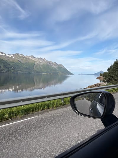 The image depicts a scenic view from a car traveling along a road near Neverdal, Norway. The foreground shows the asphalt road and a portion of the car's interior, including the side mirror which reflects the road behind the vehicle. A silver guardrail runs along the side of the road, separating it from a grassy verge. In the mid-ground, a calm body of water, likely a fjord, stretches out, reflecting the sky and the mountainous landscape. The mountains in the background are covered in green vegetation at the lower slopes and have patches of snow or white rock near their peaks. The sky is blue with scattered white clouds. The overall impression is of a serene, natural landscape viewed from a moving vehicle.