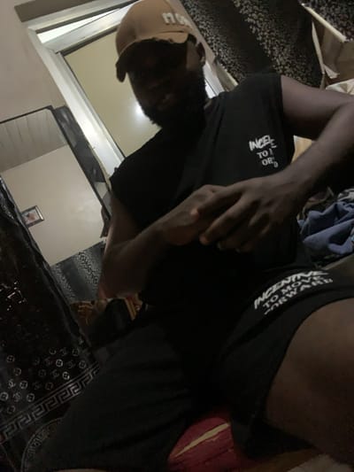 A low-angle shot depicts a person indoors. They're wearing a tan baseball cap, a black t-shirt with white text, and dark shorts. The text on the shirt appears to read "Incentive to move forward". The person's face is partially obscured. In the background, there's a window, a wall, and a patterned rug. A framed picture is also visible on the wall. The lighting is dim, creating shadows.