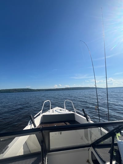 The image shows a view from the perspective of someone inside a boat on a body of water. The boat has white railings and a windshield. Two fishing rods are prominently placed on the right side of the boat, with fishing line visible. The water is a deep blue and appears to be slightly choppy. In the distance, a tree-covered shoreline is visible under a bright blue sky with scattered white clouds and the sun shining from the upper right, creating lens flare. The scene is consistent with a location on a lake or a large river in Siukterka, Russia, likely during daylight hours.