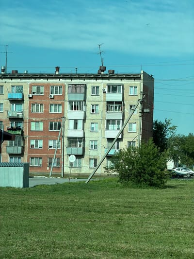 The image depicts a multi-story apartment building in Yevsino, Russia, under a bright, clear blue sky with a few wispy clouds. The building, likely from the Soviet era, features a faded, textured facade with alternating sections of light beige and reddish-brown. Numerous windows, many with white frames, punctuate the building's exterior, some with balconies enclosed with glass or featuring railings. Air conditioning units are visible mounted below some windows.
The building's roof is flat and appears somewhat weathered, with several metal chimneys and ventilation pipes protruding from it, alongside antennas. Utility wires crisscross the sky above the building. A large, concrete utility pole runs diagonally across the facade, supporting a network of electrical cables and what appears to be a transformer box. A circular sign is affixed to this pole.
In the foreground, a well-maintained lawn of green grass stretches out, appearing slightly uneven with some areas of dryer, lighter-colored grass. To the left of the apartment building, a small, metallic shed-like structure with ribbed siding is visible. Behind the building and to the right, a cluster of lush green trees provides a contrast to the urban architecture. A light-colored car and a darker car are parked on a paved area behind the trees. The overall scene suggests a typical residential area in a Russian town.
