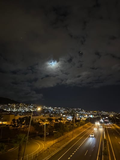 The image depicts a nocturnal urban landscape in Adeje, Spain, characterized by a highway cutting through the scene and a sprawling cityscape illuminated by artificial lights. A bright moon is visible in the upper portion of the sky, partially obscured by clouds, casting a soft glow over the scene.
The foreground features a multi-lane highway with vehicles in motion, their headlights and taillights creating streaks of light. The highway is flanked by a barrier and streetlights. To the left of the highway, there is a curved road with a streetlight and some vegetation, including palm trees.
In the middle ground, there are buildings with illuminated windows and structures, suggesting a residential or commercial area. The city extends towards the background, with lights climbing a hillside, indicating its presence on hilly terrain. The overall impression is that of a vibrant city at night, with the highway serving as a central artery.