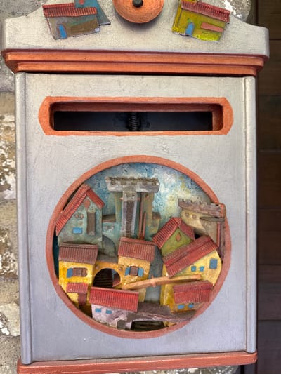 This is a close-up, eye-level, outdoor, natural light photograph of a decorative mailbox with a miniature village scene. The mailbox is primarily silver with copper-colored trim. The village scene is circular and protrudes from the front of the mailbox, depicting a cluster of small, colorful buildings with red tile roofs, reminiscent of the architecture in Dolceacqua, Italy. A central castle-like structure dominates the background of the miniature scene. The mailbox has a horizontal mail slot above the village. The background consists of a textured wall on the left and wooden planks on the right.