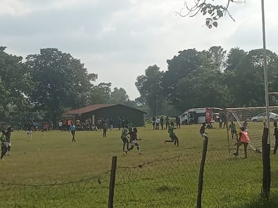 The scene depicts an outdoor football match or practice session in the Isukha North ward, Kenya, under an overcast sky. In the immediate foreground, a barbed wire fence with wooden posts stretches across the frame. On the grassy field, approximately a dozen male individuals are actively engaged in play. Players are primarily clad in green and black kits; one player near the right goal is visible in an orange bib, and another individual in a light blue shirt, likely an official, walks near the center of the field. A white football goal with a net is situated on the right side of the field. In the mid-ground, a crowd of approximately 30-40 spectators, a mix of standing and seated individuals, observes the event near a building with a dark brown corrugated roof and dark walls. To the right of the building, a white ambulance with prominent red emergency markings is parked, along with a white passenger vehicle. The background is characterized by dense green foliage and numerous tall trees. Overhanging branches from an unseen tree are partially visible in the upper right portion of the image.