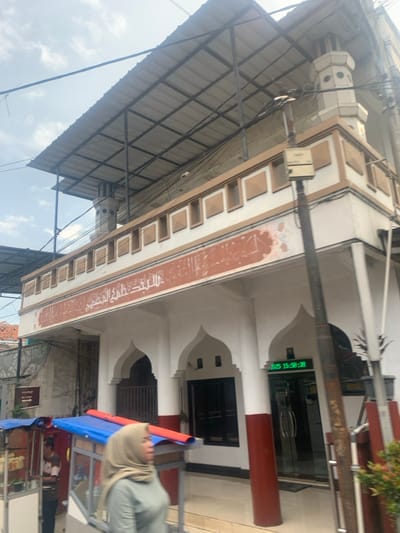 The image shows the front entrance of a mosque located at latitude -6.922, longitude 107.539 in the city of Lagadar, Indonesia. It's a white building with a red awning above the entrance and red pillars on either side. A sign above the door reads "Masjid Al-Ikhlas". The entrance has two large arched doors. On the left side of the image, there is a person standing near a cart. A woman wearing a green shirt and a hijab is walking past the cart. There is a small plant in a pot on the right side of the image. There is a digital clock on the wall that displays the time 15:58:28. The mosque is situated in a densely populated residential area, with several nearby houses. The sky is cloudy. Several electrical wires run across the image. The image is taken from a low angle, looking upward at the mosque.