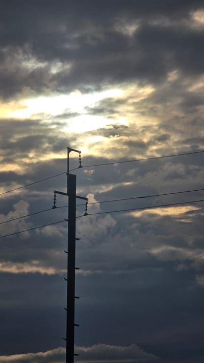 The image displays a tall, dark utility pole, likely in Leninogorsk, Russia, standing in stark silhouette against a heavily clouded sky. The pole, positioned on the left side of the frame and extending from the bottom upwards, features multiple cross arms or brackets from which bell-shaped insulators are visible, supporting several taut power lines that stretch horizontally across the middle ground. Smaller, ladder-like attachments are present on the right side of the pole. The sky is dominated by dense, dark grey and blue-grey clouds, yet a brilliant, golden-white light source, consistent with a partially obscured sun, radiates intensely from the upper-left to the central part of the frame, illuminating the edges of some clouds with a golden hue and creating significant contrast. No individuals, animals, or explicit actions are depicted within the scene.