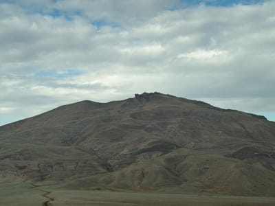 The image shows a wide, undulating mountain range under a cloudy sky. The landscape is predominantly shades of brown and green, with the mountain slopes textured by numerous gullies and ridges. The peaks of the mountain range are rugged, with some rocky outcrops visible at the highest point. In the foreground, a flat, grassy plain stretches towards the base of the mountains, with a faint, winding dry creek bed visible. The sky is filled with a mix of white and gray clouds, with patches of blue sky peeking through. The overall impression is one of a vast, natural landscape, possibly a remote or sparsely populated area. The image does not depict any people, specific objects, or actions. Given the prompt's request for location in Jeyrankechmaz, Azerbaijan, this scene is consistent with the mountainous terrain found in parts of Azerbaijan.
