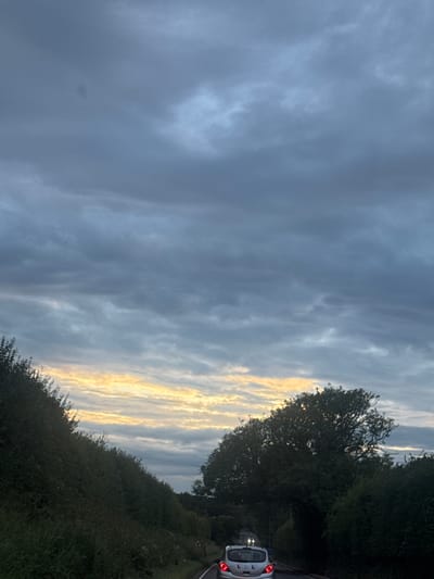 The image depicts a cloudy sky with a sliver of golden light breaking through the horizon, suggesting either dawn or dusk. A dense green hedge lines the left side of a road, which curves into the distance. On the right side of the road, large trees obscure the view. A white car, with its rear brake lights illuminated, is traveling away from the viewer on the road. The scene appears to be a rural or semi-rural setting in the Borough of Swale, United Kingdom. There are no people visible in the image. The overall atmosphere is somber due to the heavy cloud cover, but the glimpse of sunlight adds a touch of warmth.