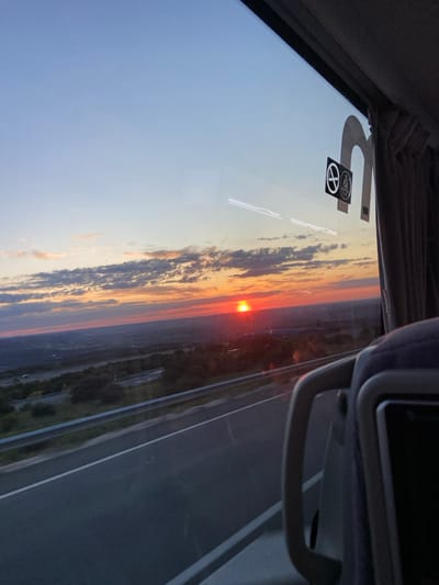 The image shows the sunset over a highway, viewed from inside a vehicle. The vehicle is moving, as indicated by the blurred road and trees. The sun is setting in the distance, casting an orange glow over the sky and surrounding landscape. The highway is a four-lane road with a white dividing line. The location is Otero de Herreros, Spain, based on the provided coordinates. The scene is captured through a window, with a partial view of the interior of the vehicle, including a seat and a curtain. A no-smoking sticker is visible on the window.