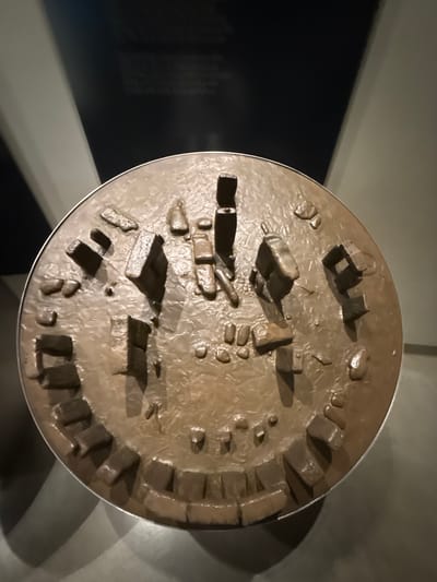 A circular, bronze-toned scale model of the prehistoric monument of Stonehenge is displayed, positioned within a visitor center near Amesbury, United Kingdom. The model features numerous raised elements representing the sarsen stones and bluestones, with some standing vertically and others lying horizontally, arranged in concentric circular and horseshoe patterns on a textured base. An outer ring composed of smaller, rectangular blocks encircles the primary stone arrangements. The model rests on an unseen display surface. The background consists of a dark, indeterminate wall, exhibiting subtle reflections and faint, indistinct textual information, consistent with an indoor museum or exhibition environment. Direct illumination from above casts distinct shadows from the model's raised features onto its textured surface. No individuals are present in the scene.
