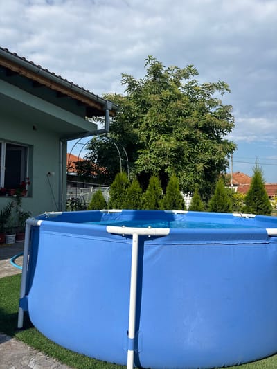 The image depicts an outdoor scene, likely a backyard, in Dalgopol, Bulgaria. In the foreground, a large, blue, above-ground swimming pool with white support poles is prominently featured. The pool appears to be filled with water and is covered with a solar cover. The pool is situated on a patch of artificial turf, which is bordered by a stone pathway.
To the left of the pool, a light green house is visible. The house has a window with red flowers in pots on the windowsill and a gutter system running along its roof. A black electrical cord extends from an outlet on the exterior wall of the house.
Behind the pool and house, a dense green tree with white blossoms or small fruits dominates the background. Several conical evergreen trees are planted in a row in front of the larger tree. In the distance, the roofs of other houses with red tiles are visible. The sky is partly cloudy. The scene suggests a residential area on a bright, possibly warm day.