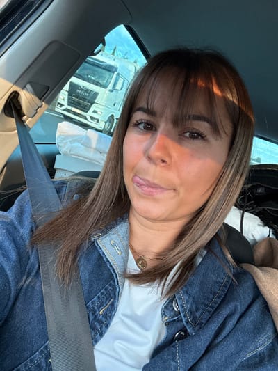 This image depicts a scene in Bourg-lès-Valence, France, captured from within a moving vehicle. A female individual, approximately in her 20s or 30s, occupies the front passenger seat, identifiable by the gray seatbelt crossing her torso diagonally from upper left to lower right. She has medium-brown hair with bangs, a tanned complexion, and is wearing a blue denim jacket over a white t-shirt, accessorized with a gold-colored chain necklace featuring a circular pendant. Her gaze is directed at the camera, with her tongue slightly protruding from the left corner of her mouth, indicating a deliberate pose. Through the vehicle's front passenger window, a white MAN brand semi-truck is visible on a multi-lane highway, traveling in daylight under a blue sky with some cloud cover. Distant vegetation is discernible along the highway's edge. The vehicle's interior features gray trim on the A-pillar and roof, and items such as a white package and a brown textile are present in the rear seat.